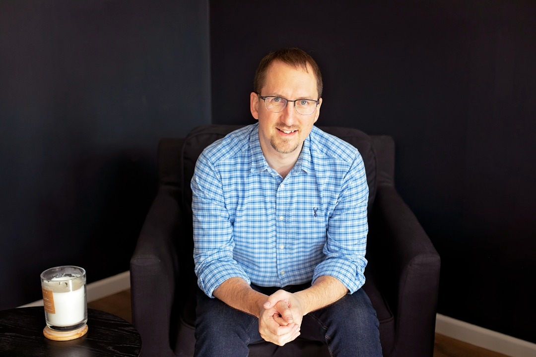 Mike sitting, looking at the camera in a dark-coloured room next to a coffee table with a candle on it.